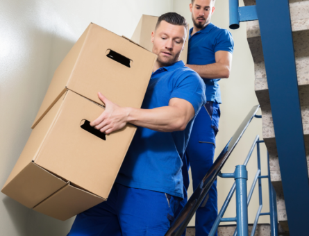 Two men with blue uniforms moving boxes down stairs